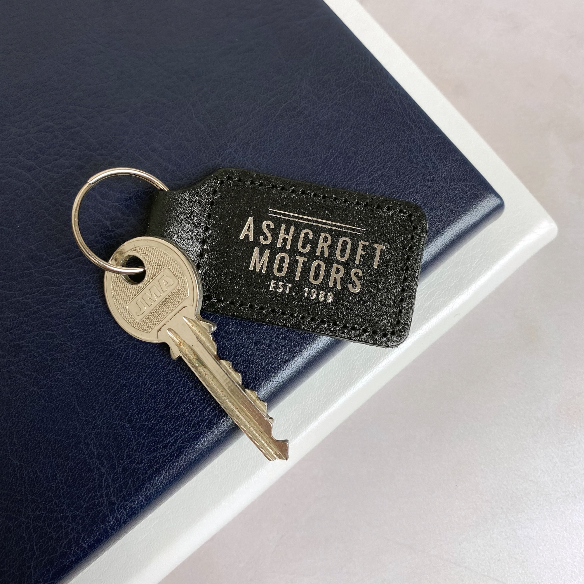 Branded black leather keyring with silver business logo, silver split ring, and a key attached.